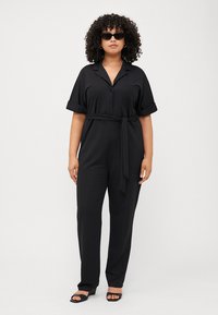 A woman with curly black hair wears a black belted jumpsuit and sunglasses, standing against a neutral background.