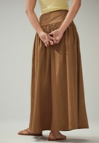Jupe en coton marron, longueur complète, avec une taille froncée. Le modèle se tient debout, les mains jointes derrière, portant des sandales plates.