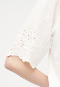 Λευκό κοντομάνικο με κεντήματα με άνθη και περίγραμμα με κυματιστό σχήμα στον βραχίονα ενός ατόμου.