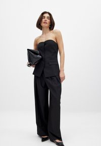 Musta olkaimeton yläosa napilla, yhdistettynä leveälahkeisiin mustiin housuihin. Pitelee mustaa taitettavaa laukkua. Sileä kangas ja räätälöity istuvuus.