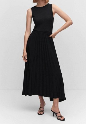 Femme portant une robe maxi noire sans manches avec une jupe plissée et des sandales noires à talons hauts à brides, debout avec une main sur la taille.