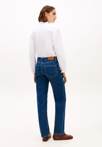 Μπλε τζιν παντελόνι με ίσια γραμμή, που έχει ένα δερμάτινο λογότυπο της μάρκας στο πίσω μέρος, φορεμένο με λευκό πουκάμισο και καφέ παπούτσια.