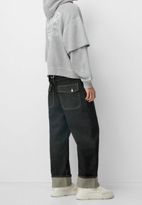 Felpa grigia con dettaglio del logo, abbinata a jeans neri a gamba larga con orlo risvoltato, completata da sneakers bianche. Stile casual streetwear.