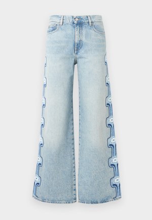 Nő kék, rövid ujjú mintás ingben, világos, bő szárú farmernadrágban oldalán virágmintával, barna övvel és fekete magas sarkú szandálban áll.