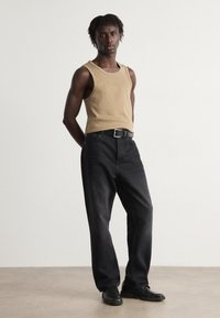 Top din bumbac bej, fără mâneci, cu decolteu larg, purtat cu pantaloni din denim negri. Curea neagră cu cataramă argintie, asortată cu pantofi negri.