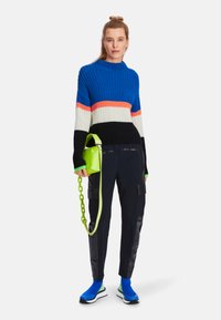 Strickpullover mit blauen, orangefarbenen, weißen und schwarzen Streifen; schwarze Hose mit Lederelementen; neongrüner Handtasche mit Kettenriemen; blaue Sneakers.
