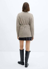 Cardigan en maille côtelée beige avec une ceinture à la taille, doté de manches longues et d'une coupe décontractée, associé à une jupe plissée noire et des bottes mi-hautes.