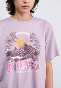 Lilac cotton t-shirt featuring a mountain graphic, sun, and floral accents, with “ROXY” in bold pink letters at the center.