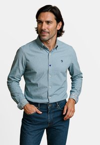 Lichtblauwe blouse met een subtiel patroon, contrasterend kraagontwerp en witte knopen, gecombineerd met donkere denim jeans.