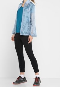 Kvinna iklädd en ljusblå vindjacka, teal-färgad ärmlös topp, svarta leggings, vita strumpor och svarta trailskor med röda sulor och snören.