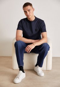Tricou navy cu mânecă scurtă, dotat cu o etichetă logo pătrată, asortat cu blugi denim albastri și adidași albi, așezat pe un scaun texturat de culoare bej.