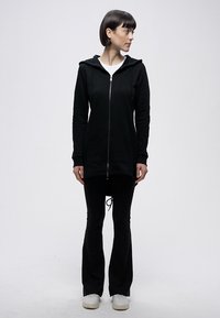 Fekete cipzáras kapucnis pulóver kapucnival, hosszú ujjakkal és elülső zsebekkel, fekete bővülő szárú nadrággal és fehér tornacipővel párosítva. Sima szövet textúra.