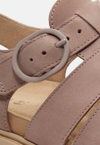 Close-up van een taupe suède sandaalriem met een ronde kunststof gesp en zichtbare stiksels op een beige gevoerde voetbed.