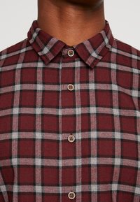 Chemise à carreaux bordeaux, noire et grise avec boutons et col plié portée par une personne à la peau foncée.