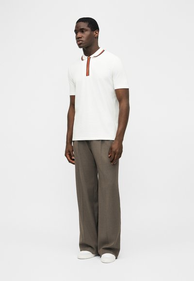 Άνδρας που στέκεται και φοράει λευκό πόλο με κοντά μανίκια και πορτοκαλί λεπτομέρειες, φαρδιά γκρι παντελόνια και λευκά αθλητικά παπούτσια, μπροστά σε απλό ανοιχτό φόντο.