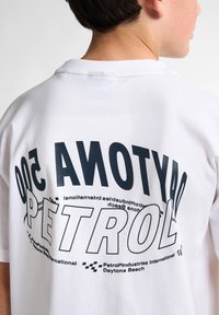 Bílá bavlněná trička s velkým černým potiskem na zádech, zobrazující "DAYTONA 500" a "PETROL" tučným písmem.