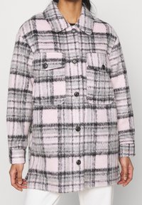 Chaqueta de franela de gran tamaño en cuadros grises y rosas, con cuello tipo camisa, cierre delantero de botones y dos bolsillos en el pecho. Textura suave.
