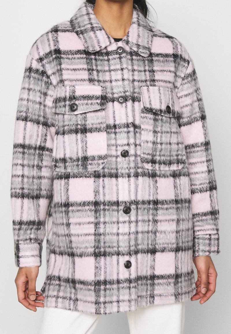 Chaqueta de franela de gran tamaño en cuadros grises y rosas, con cuello tipo camisa, cierre delantero de botones y dos bolsillos en el pecho. Textura suave.