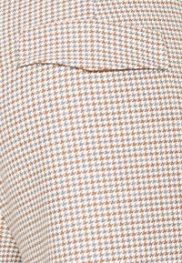 Houndstooth-mönstrad textil i vitt, grått och rostorange med synlig fickdetalj, som har en texturerad vävd look.