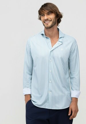 Hombre sonriente con cabello castaño medio que lleva una camisa de botones a rayas azul claro y blanca y pantalones oscuros, de pie con una mano en el bolsillo.