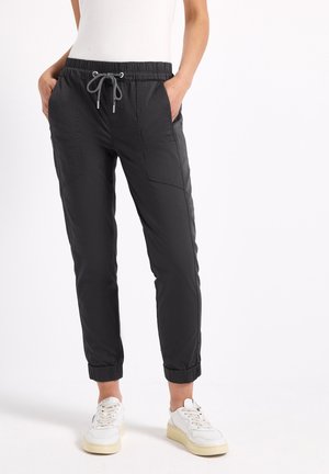 Person wearing black drawstring jogger pants with front pockets and white sneakers, standing with hands in pockets against white background.