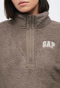 Szara bluza polarowa z suwakiem do ćwiartki. Charakteryzuje się wypukłą fakturą, haftowanym białym logo „GAP” oraz wysokim kołnierzem.