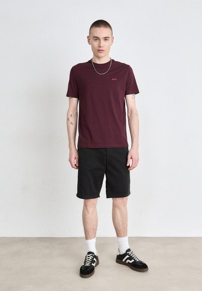 T-shirt en coton bourgogne avec logo rouge, associé à un short noir, des chaussettes blanches montantes et des baskets noires avec des accents blancs. Le modèle se tient devant un fond neutre.