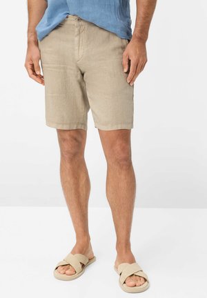 Männliches Model trägt beige knielange Shorts, blaues Hemd und beige Sandalen mit Kreuzriemen und steht vor weißem Hintergrund.