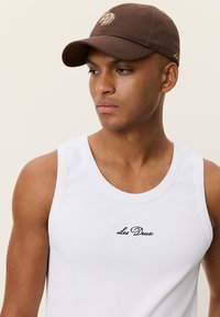 Débardeur blanc côtelé avec "Les Deux" brodé en noir, associé à une casquette de baseball marron ornée d'un logo.