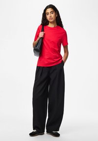 Rød bomulds t-shirt med rund hals, kombineret med løse sorte bukser. Modellen holder en sort skuldertaske og har sorte sneakers på.