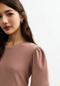 Haut rose texturé à manches longues, avec un col rond et des manches bouffantes. Le tissu présente un motif subtil, renforçant son apparence dimensionnelle.