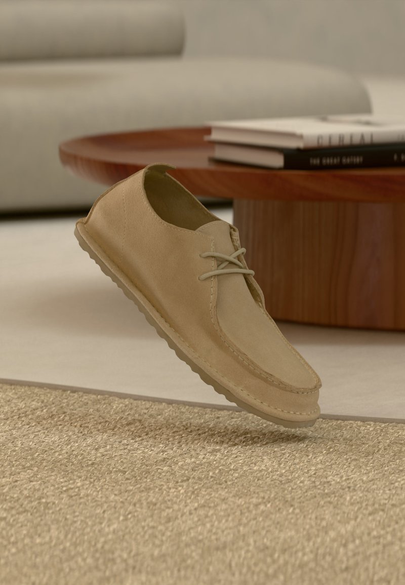 Chaussure décontractée à lacets en daim beige flottant au-dessus d'un tapis texturé dans un salon moderne avec une table basse en bois et des livres.
