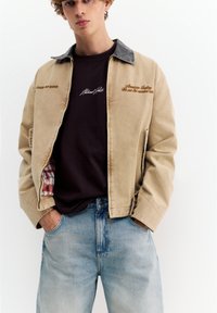 Ung mand iført beige jakke med brun tekst, sort skjorte og lyseblå jeans, hænderne i lommerne, mod hvid baggrund.