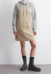 Beige cordfluweled overall klänning med justerbara band, matchad med en grå hoodie och gröna kraftiga ankelstövlar. Slät textur, avslappnad design.