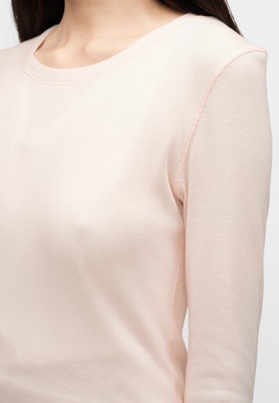 Γυναίκα που φοράει ροζ ανοιχτό ροζ μπλούζα με μακριά μανίκια και στρογγυλή λαιμόκοψη, εμφανιζόμενη από τους ώμους μέχρι τη μέση, πάνω σε απλό φόντο.