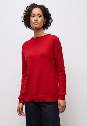 Frau mit kurzen lockigen Haaren, die einen einfarbigen roten Langarmpullover und dunkelblaue Jeans trägt, stehend vor einem schlichten hellen Hintergrund.