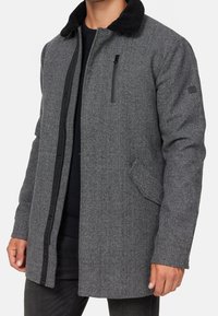 Abrigo gris de espiga con cuello de sherpa negro, cierre frontal de cremallera y botones a presión, bolsillos laterales y un bolsillo en el pecho con cremallera.