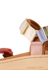 Sandal dengan tali yang menampilkan tali pastel krem dengan perangkat keras emas, bantalan kaki suede, dan sol karet alami yang diberi label "NATURAL SOFT™."