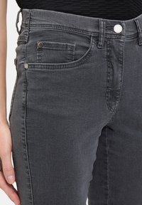 Le jean en denim gris présente un design ajusté avec une taille mi-haute, des poches avant et des accents en métal. La texture semble lisse et légèrement décolorée.