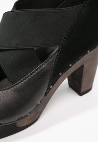 Chaussure noire à talon avec une tige en cuir texturé, une sangle élastique et un talon en bois. Présente des accents métalliques le long de la couture.