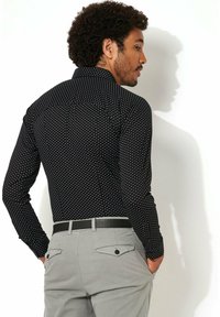 Zwart overhemd met een witte geometrische print, standaard kraag, gedragen met lichtgrijze broek en een zwarte riem. Achteraanzicht.