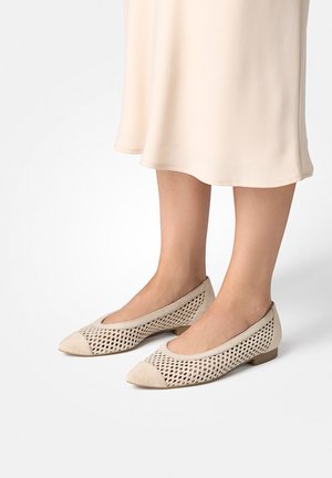 Beige perforierte Schuhe mit niedrigem Absatz, getragen zu einem hellcremefarbenen Midirock, vor weißem Hintergrund.