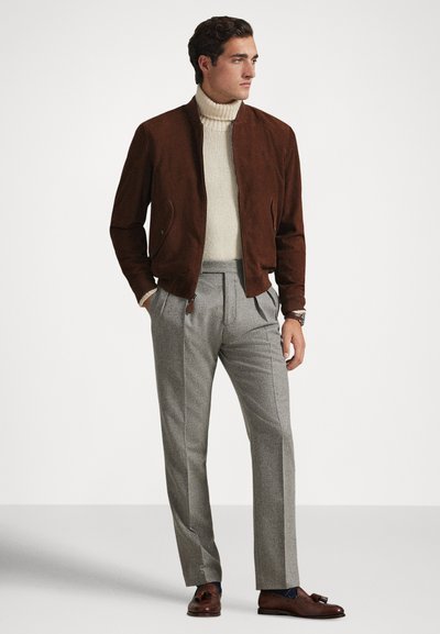 Moški, oblečen v rjavo jakno z zadrgo, kremni rolkasti pulover, sive nagubane hlače in rjave usnjene cipele z resami, stoji z rokami v žepih.