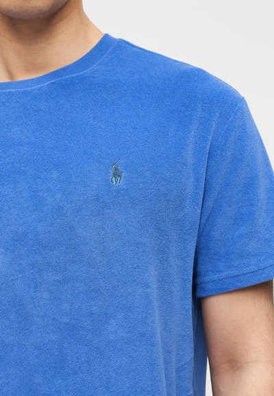 Άνδρας που φοράει μπλε πουκάμισο από πετσέτα με κοντά μανίκια και ένα μικρό σκούρο μπλε λογότυπο παίκτη πόλο στο στήθος.