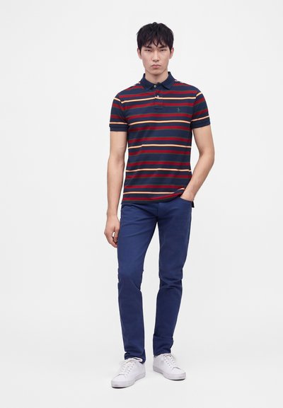 Νέος άνδρας που φοράει μπλε μπλουζάκι πόλο με ρίγες, μπλε παντελόνι και λευκά αθλητικά παπούτσια, στέκεται με το ένα χέρι στην τσέπη μπροστά σε λευκό φόντο.