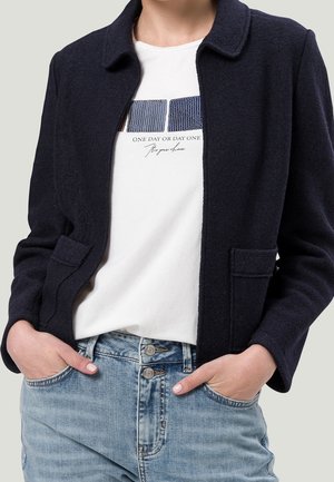 Persoon draagt een marineblauwe jas over een wit T-shirt met blauwe vierkante patronen en tekst, gecombineerd met lichtblauwe high-waisted jeans, handen in de zakken.