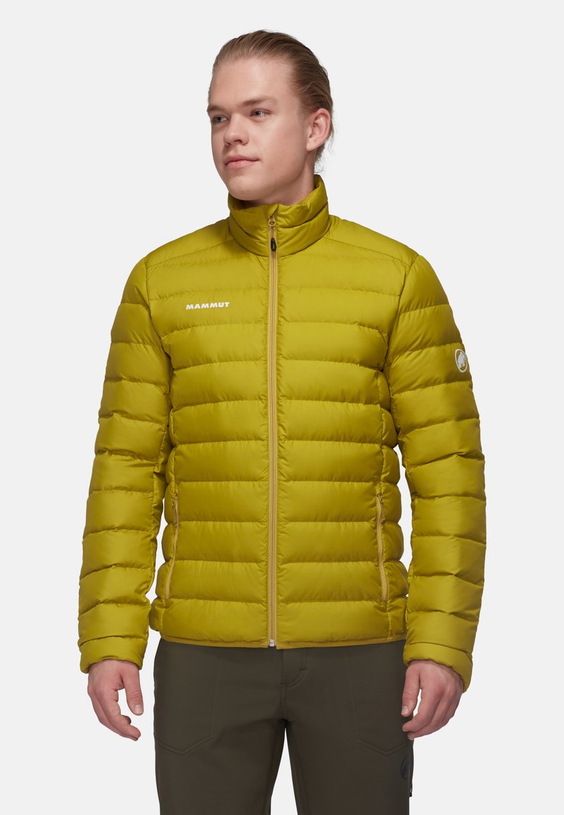 Kurtka puchowa w jasnym oliwkowym kolorze z pikowanym wzorem, wysokim kołnierzem, zamkiem z przodu i logo Mammut po lewej stronie klatki piersiowej. Miękka tekstura.