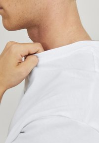 Hvid bomulds t-shirt med ribbet halskant, der bliver blidt grebet om kraven. Fokuser på stofets tekstur og designklarhed.