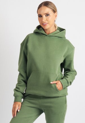 Zelená mikina s kapucňou z mäkkého materiálu s veľkou prednou kapsou, voľným strihom a nastaviteľnou šnúrkou na kapucni, v kombinácii s rovnakými nohavicami.