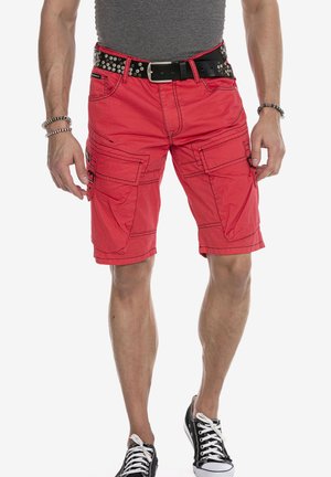 Rote Cargo-Shorts mit mehreren Taschen, Kontrastnähten und einem schwarzen Nieten-Gürtel. Getragen mit einem grauen, enganliegenden Shirt und schwarzen Sneakers.